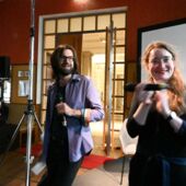 Andreas Scherffig and Hanna Hamel stand in front of a screen and hold microphones in their hands. Hanna Hamel claps her hands.