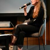 Hanna Hamel sitzt auf einem Stuhl auf einem Podium und spricht in ein Mikrofon. Sie ist im Profil zu sehen.