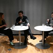 Pola Groß, Jona Spreter and Ivana Sokola are sitting at two round bistro tables in front of a gray curtain. There are glasses of water in front of them and they are facing each other in conversation.