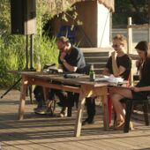 Cord Riechelmann, Moritz Gansen and Tabea Hertzog sit at a table made of chipboard on the open-air stage at Floating Berlin. Tabea Hertzog reads, the other two are listening to her.