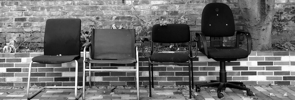 Schwarzweißfoto: Vor einer Ziegelmauer stehen vier verschiedene Stühle, von links nach rechts: ein geschwungener Stuhl ohne Armlehnen, ein Freischwinger, ein Stuhl mit unten ausgesparter Rückenlehne und Armlehnen, ein Bürostuhl mit Rollen.