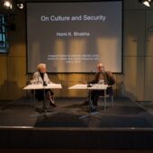 Sigrid Weigel and Homi Bhabha are sitting at two tables on a stage, a presentation “On Culture and Security” is running in the background