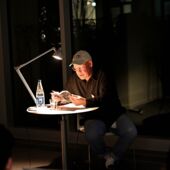 Tim Holland sits at a small round table, illuminated by a desk lamp, and reads from a book.