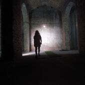 A very dark room, in the background a written wall illuminated with a lamp. In the middle of the darkness runs the silhouette of a woman.