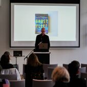 Anders Engberg-Pedersen stands at the lectern in front of a screen and speaks. The cover of Peter Singer's "Ghost Fleet" can be seen on the screen.