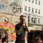 Moritz Gansen stands on the pedestal of the “Dancing Couple” statue on Hermannplatz in Berlin-Neukölln and speaks into a microphone. Posters for a rally can be seen behind him, with a McDonald's joint in the background on the right.
