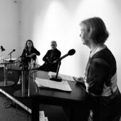 Black and white photograph of Mona Körte, Stefan Willer and Ulrike Vedder. Ulrike Vedder stands in the foreground and speaks into a microphone at the lectern. Stefan Willer and Mona Körte sit in the background, watching Ulrike Vedder.