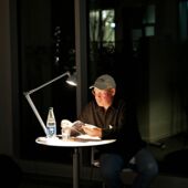 Tim Holland sits at a small round table, illuminated by a desk lamp, and reads from a book.