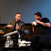 Der Autor Hannes Bajohr und der Moderator Morten Paul sitzen vor einer großen Leinwand und unterhalten sich.