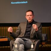 The author Hannes Bajohr is sitting on a podium in front of a screen. On this screen you can read the words “Sequenziell vs. Konnektionistisch.”