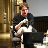 Der Autor Elias Hirschl sitzt an einem Laptop und spricht in ein Mikrofon.
