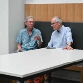 John Rieder and Axel Goodbody sit on an upholstered bench at a conference table and talk.