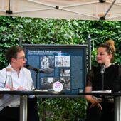Eva Geulen and Teresa Präauer are sitting on a stage and talking. Behind them is an information board about the garden at the Literaturhaus.