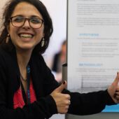 Marwa El Chab stands in front of a poster, gives two thumbs up and smiles