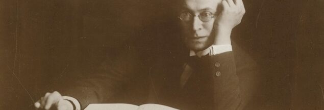 Photo of Karl Kraus. He sits at the table in a suit and glasses and looks into the camera. On the table lies an open book and a closed notebook next to it. Kraus rests one elbow on the table, his hand on his temple.
