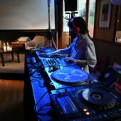 The DJ Mo Chan / DJ Kohlrabi is standing at a mixing desk bathed in blue light. She wears a red headset.
