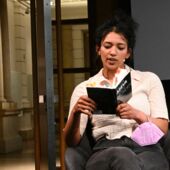 Elisa Aseva sitzt auf einem Stuhl und liest aus ihrem Buch »Über Stunden«, in dem bunte Klebezettel kleben. Am Handgelenk trägt sie eine violette FFP2-Maske.