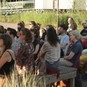 Circa 25 Menschen sitzen im Publikum auf der Freilichtbühne von Floating Berlin und hören der Lesung zu.