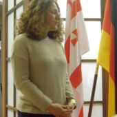 A woman stands in front of a Georgian, a German and a European flag.