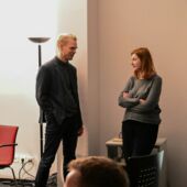 Matthias Schwartz and Olga Grjasnowa are talking facing each other. Schwartz is standing in front of a black floor lamp, Gryaznova is leaning against a tech cart with her arms folded.