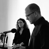 Black and white photograph of Stefan Willer and Mona Körte. In the foreground, Stefan Willer speaks into a microphone at the lectern. Monika Körte stands next to him and looks into the void.