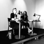 Schwarz-Weiß-Fotografie von Esther Kinsky und Mona Körte, die beiden sitzen auf einem Podium. Esther Kinsky spricht in die Menge, Mona Körte schaut sie an.