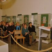 Fritz-Dieter Kupfernagel gives the speech at the exhibition opening, a well-attended audience sits in front of him in front of various exhibition showcases