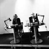 Schwarz-Weiß-Fotografie von Gertraud Klemm und Claude Haas auf einem Podium. Claude Haas spricht in ein Mikrofon, Gertraud Klemm schaut ihn an.