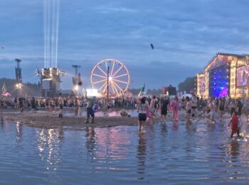 Szene vom Festival Przystanek Woodstock. Im Abendlicht ist eine große Pfütze zu sehen, in der Menschen tanzen und spielen. Hinten rechts ist eine hell beleuchtete Bühne, in der Mitte ein Riesenrad zu sehen.