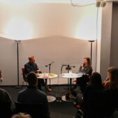 In leichter Aufsicht sind Matthias Schwartz und Olga Grjasnowa im Gespräch auf roten Stühlen sitzend vor einer weißen, durch zwei Stehlampen beleuchteten Wand zu sehen. Vor ihnen sitzt das Publikum.