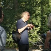 Cord Riechelmann speaks into a microphone in front of trees and a bicycle in Volkspark Hasenheide.