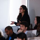 A woman in the audience stands up and asks a question, several other participants listen