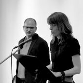 Schwarz-Weiß-Fotografie von Janika Gelinek und Stefan Willer. Janika Gelinek spricht in ein Mikrofon am Rednerpult, Stefan Willer steht neben ihr und schaut sie an.