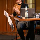 Der Autor Philipp Schönthaler sitzt auf einem Podium und spricht. Vor ihm steht ein Laptop, er wendet sich zum Publikum und hält in der rechten Hand hält er ein großes Stück Papier.