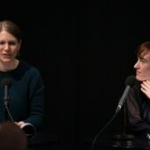 Hannah Markus speaks into a microphone in front of a black background, Nora Bossong listens to her