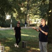 Cord Riechelmann is standing in a park under a tree and speaks into a microphone. People with headphones are standing around him listening, including Christina Ernst and Hanna Hamel.