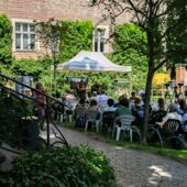 Im Garten des Literaturhauses sitzen circa 20 Personen auf Gartenstühlen, sie tragen Headsets und schauen auf ein Gartenzelt mit Bühne, auf der Janika Gelinek mit Heike Geißler spricht.