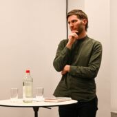 Der Preisträger Georg Simmerl steht an einem Stehtisch und schaut, die Hand am Kinn, nach rechts. Vor ihm liegt ein Buch, rechts davon stehen zwei Gläser und eine Wasserflasche.