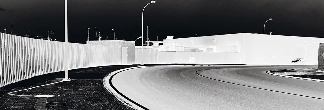 Negativbild einer scharfen Straßenkurve, die von Laternen gesäumt ist. Oben rechts ist vor einem überbeleuchteten Gebäude der Umriss eines Autos zu erkennen.