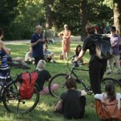 Cord Riechelmann stands on a lawn in Volkspark Hasenheide and speaks into a microphone. About 15 people with headsets sit and stand in a circle around him, some of them have bicycles with them.