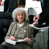 Ulrike Ottinger sitzt mit einem Buch im Schoß auf einem Stuhl und schreibt etwas hinein, neben ihr steht ein Mann