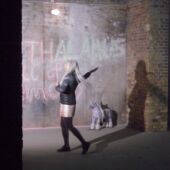 A woman in a leather jacket dances in a dark room, in front of a described concrete wall and between pillars made of bricks. In the background is a small handmade unicorn.
