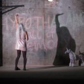A woman with long white hair and glittery top stands in front of a described concrete wall and stretches an arm in the air, next to her a small made unicorn