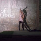 A woman with long white hair and glittery top dances in front of a concrete wall described, casting a dark shadow on the wall
