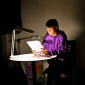 Anja Kümmel sitzt, von einer Schreibtischlampe angeleuchtet, an einem kleinen runden Tisch und liest aus einem Buch vor.