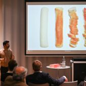 Onur Özsoy stands in front of a screen and speaks to the audience. Curry sausages in various stages of preparation can be seen on the screen.