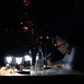 Tabea Hertzog and Cord Riechelmann sit outside at a table lit by small lanterns. Cord Riechelmann speaks into a microphone.