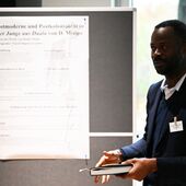 Pascal Ongossi Assamba stands in front of his poster on the topic of “Postmodernism and postcoloniality”. He holds a notebook in his hand and speaks to the audience.