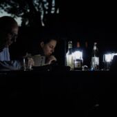 Tabea Hertzog and Cord Riechelmann sit outside at a table lit by small lanterns. Tabea Hertzog speaks into a microphone.