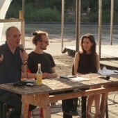 Cord Riechelmann, Moritz Gansen und Tabea Hertzog sitzen an einem Tisch aus Spanplatten auf der Freiluftbühne von Floating Berlin. Cord Riechelmann spricht ins Publikum.